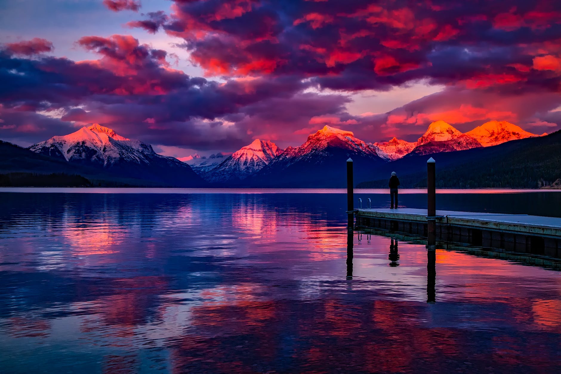 Sunset over mountains. Purple, red and orange sky. Snow-capped peaks look over a vast, still lake. A man looks at the scenic beauty on a landing near the lake.